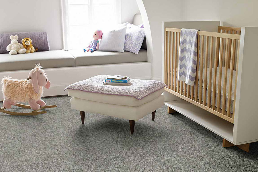 غرفة الطفل مع سجادة رمادية ومزينة بسرير حديث ، وعثماني مع بطانية طفل وردي ملفوفة فوقه ، وحصان هزاز وردي للأطفال ، ووسائد أرجوانية على نافذة جلوس.